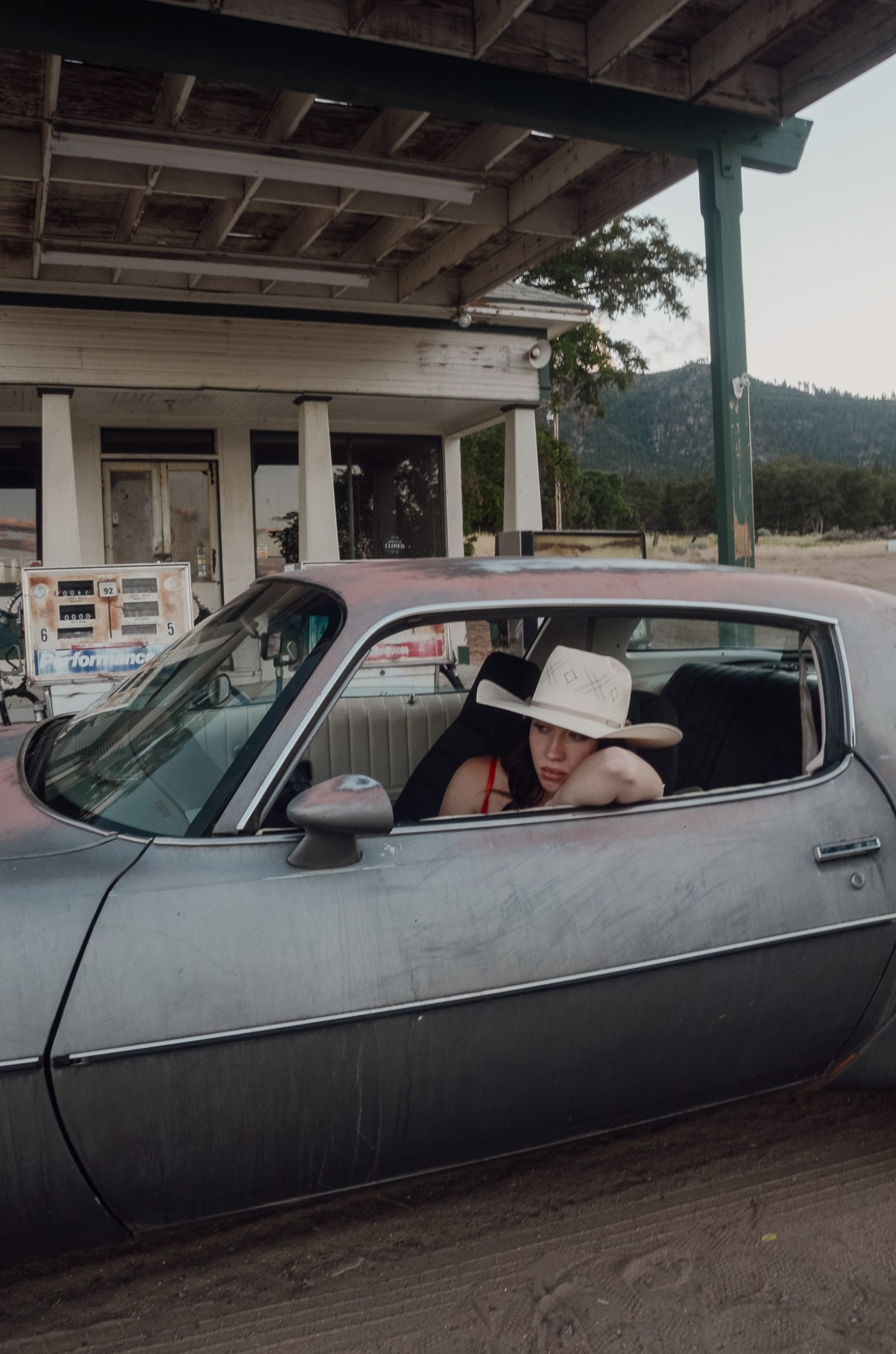 Olivia resting her arm on the window of an old car at an abandoned gas station