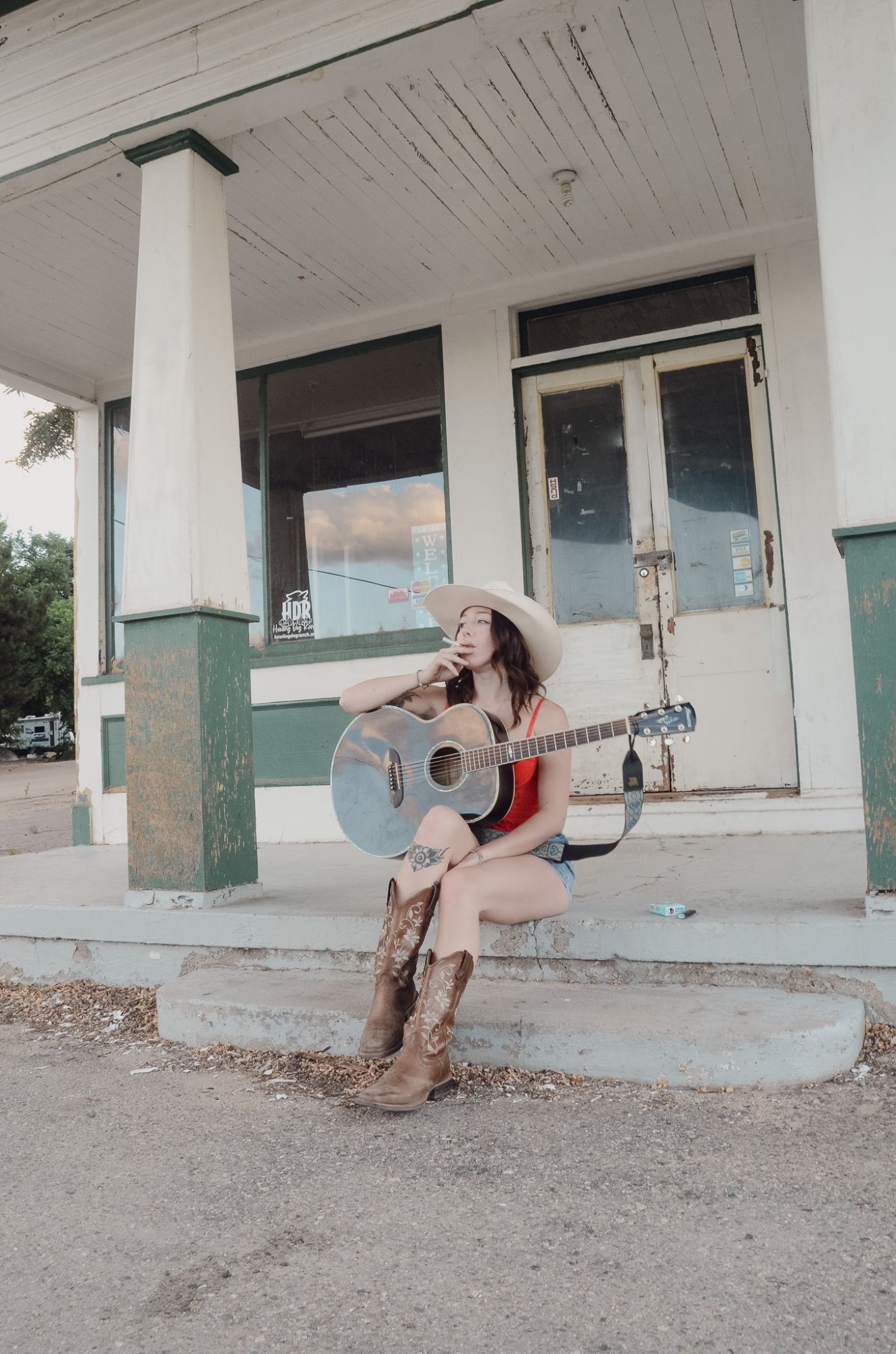 Olivia Vega sitting on the porch steps of an old general store with her acoustic guitar, in a cowboy hat and embroidered boots