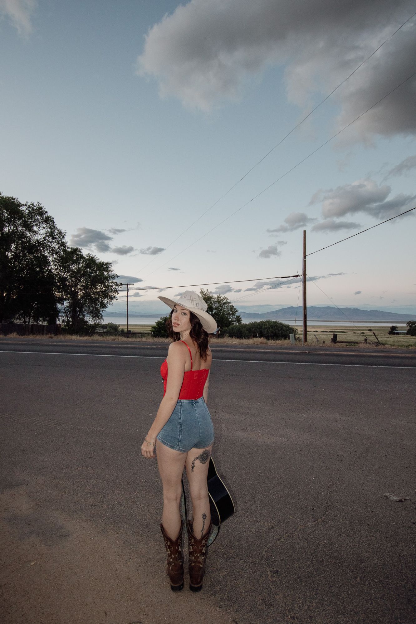 Olivia looking back over her shoulder on an empty highway at dusk, guitar in hand