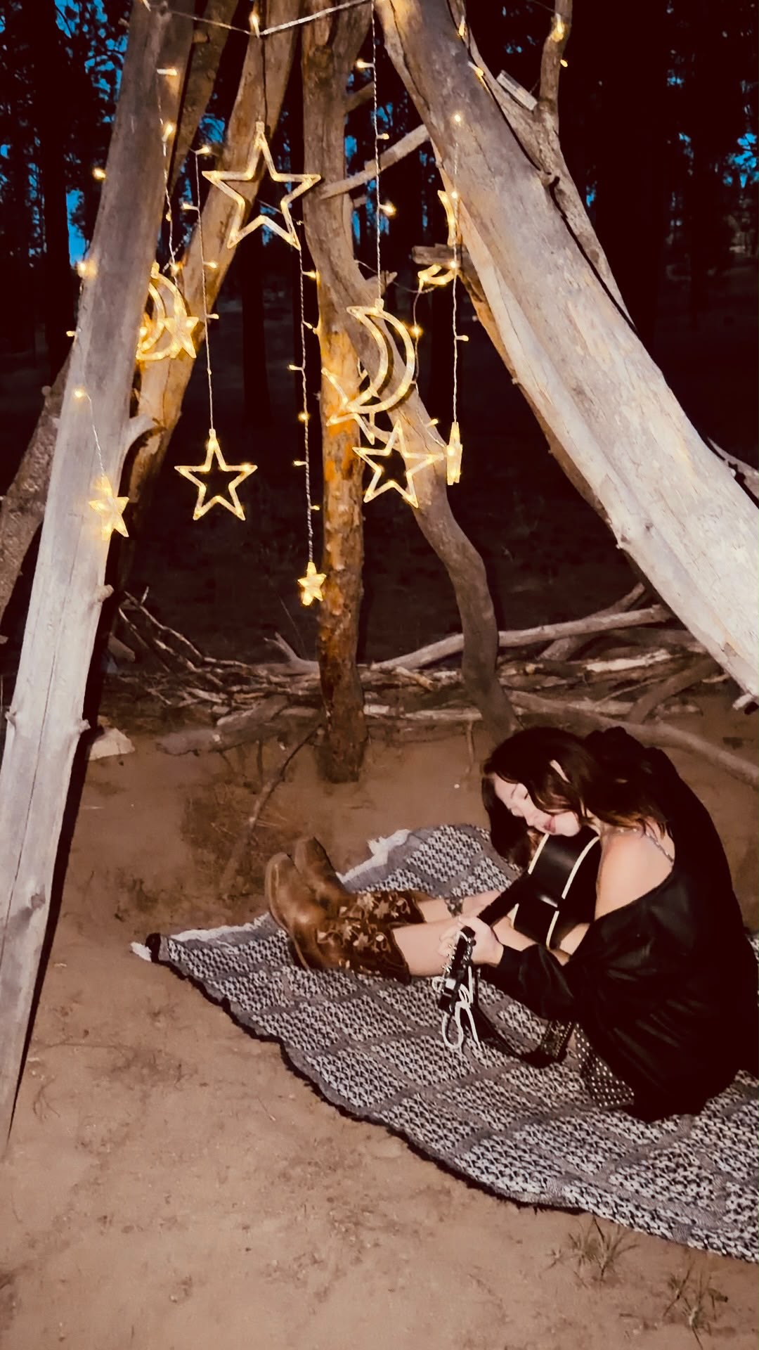 Olivia sitting with her guitar under a driftwood teepee hung with star and moon lights